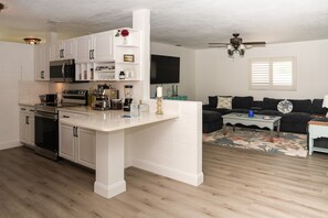 A modern kitchen with white cabinetry, a built-in microwave, and a stove. There is a dining area with a black sofa and a coffee table. The room has wooden flooring and a ceiling fan.