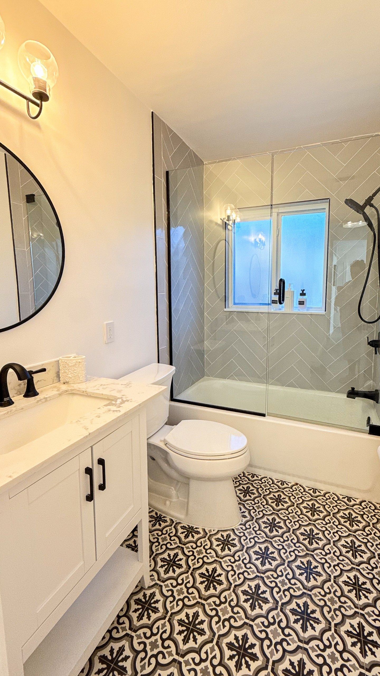 Een badkamer met een wit toilet, een badkuip, een glazen douchecabine, een ronde spiegel en een marmeren aanrechtblad.