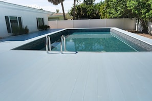 A rectangular swimming pool with a white deck, surrounded by a white fence and palm trees.