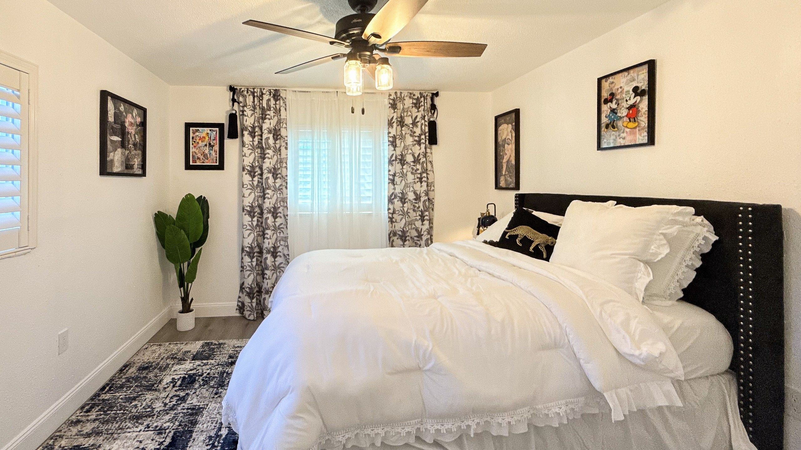A bedroom with a bed, a ceiling fan, a window with curtains, and wall-mounted pictures.