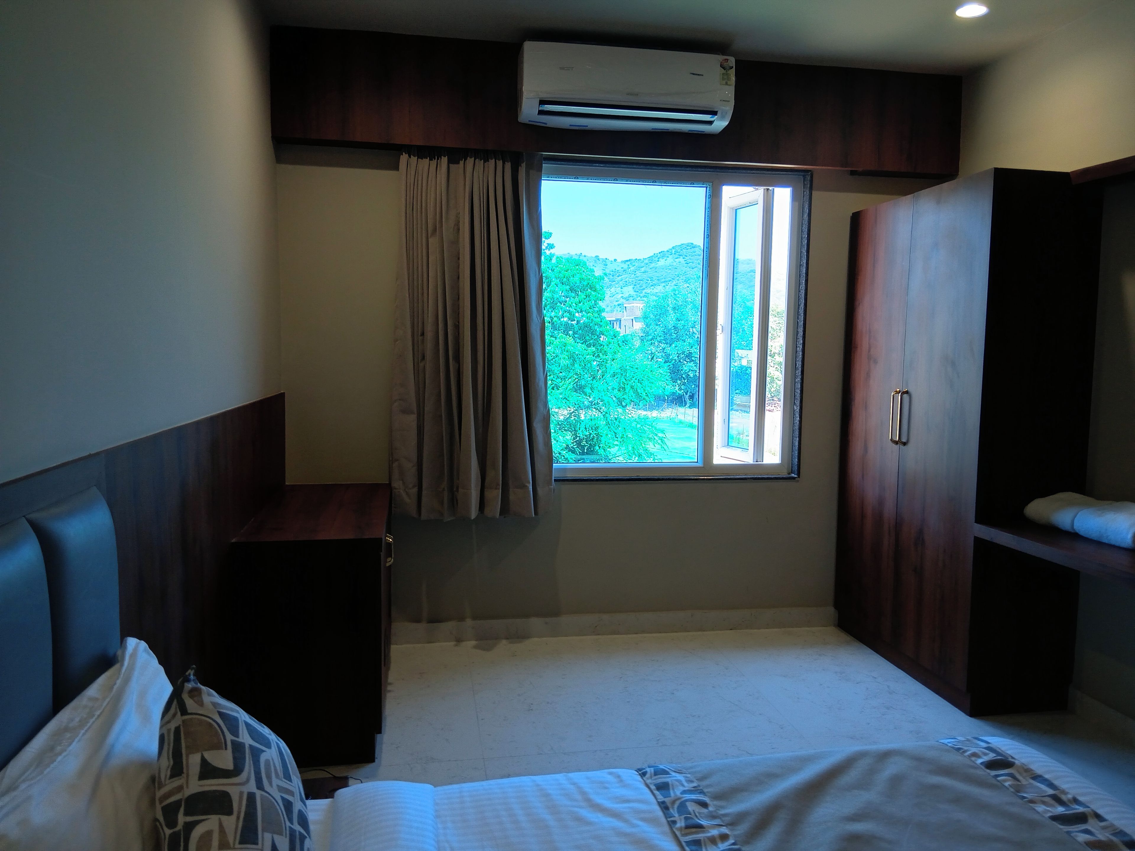 A hotel room with a bed, a desk, an air conditioning unit, and a window with curtains.