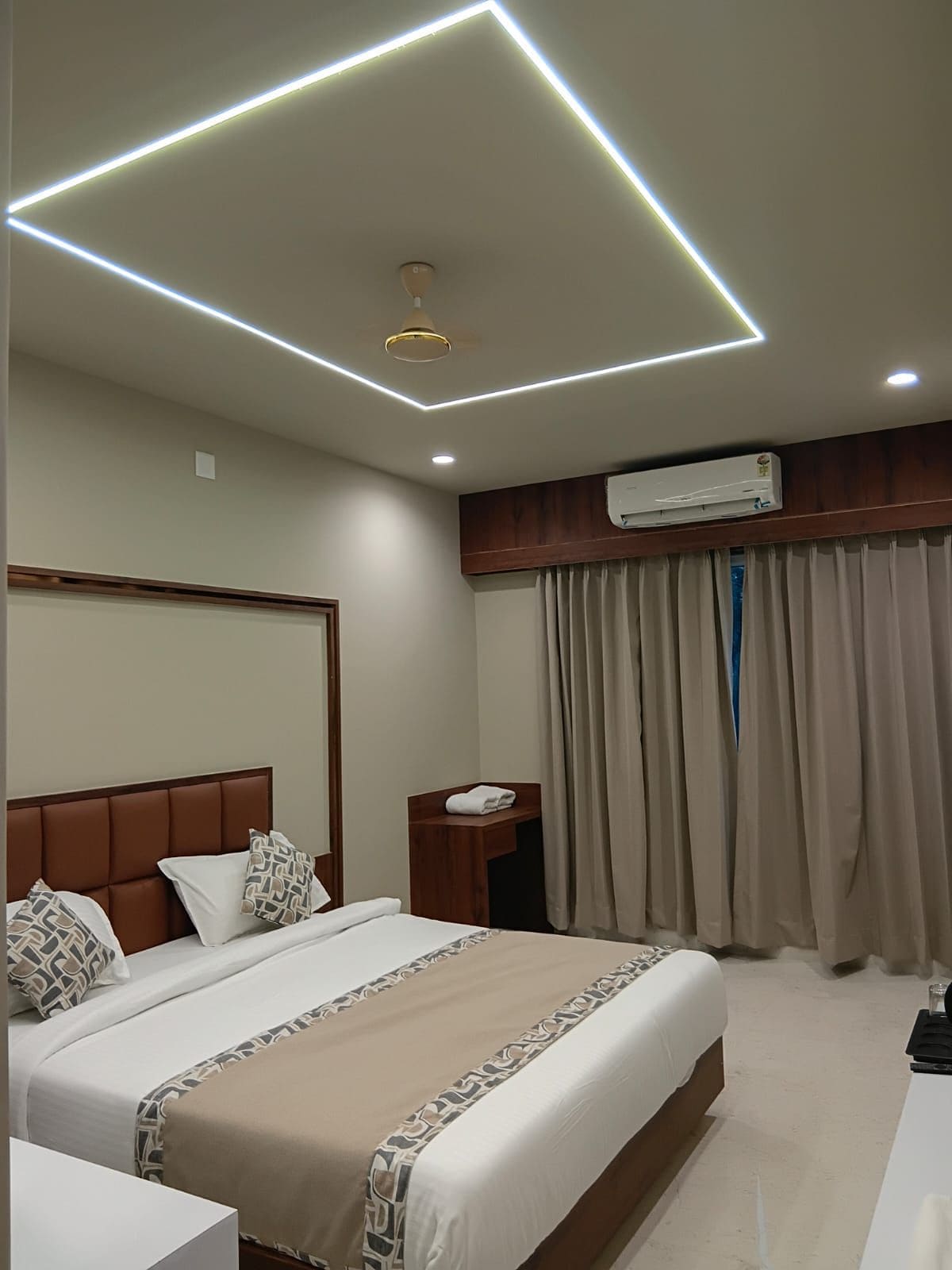 Una habitación de hotel con cama, mesita de noche, equipo de aire acondicionado y ventana con cortinas.