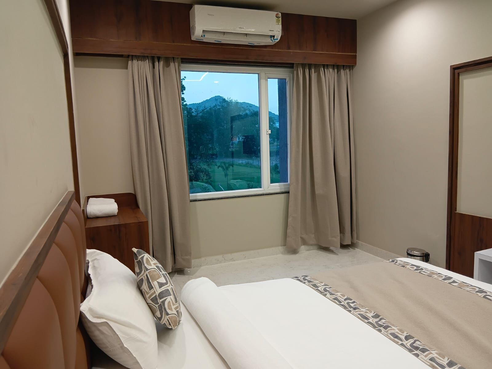 Une chambre d’hôtel avec un lit, une fenêtre avec des rideaux, un appareil de climatisation et une vue sur un paysage montagneux.