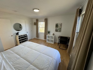 Un dormitorio con una cama grande, un aparador, un espejo, una ventana con cortinas, una mesita con una planta y una silla.