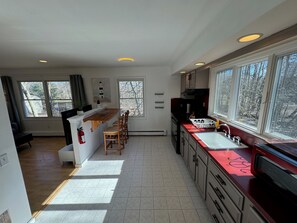 Une cuisine dotée d’un îlot central, d’un évier et d’une cuisinière. De grandes fenêtres laissent entrer la lumière naturelle et offrent une vue sur les arbres à l’extérieur.