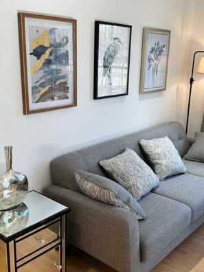 Un salon avec un canapé gris, des coussins à motifs, une table basse en verre surmontée d’un vase métallique, et trois œuvres d’art encadrées accrochées au mur.