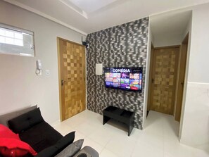 A living room with a flat-screen TV mounted on a textured wall, a sofa with cushions, and a door with a wooden pattern. - Apartment 304 · 304 2 bedrooms - Stylish and Comfortable (Belém)