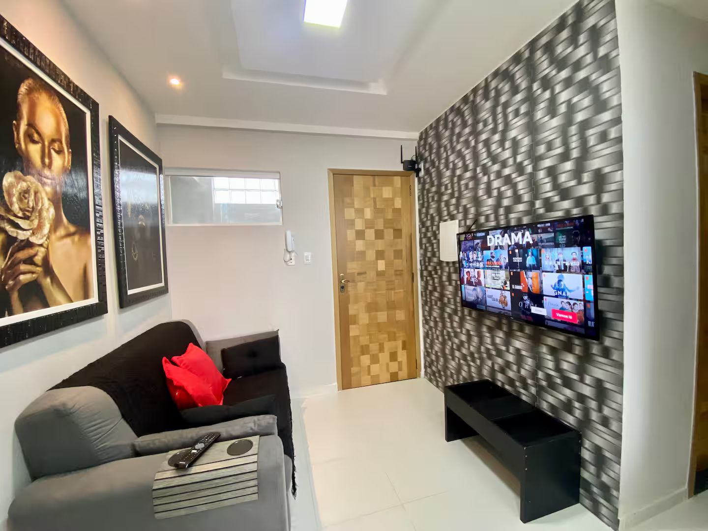 Un salon moderne avec un téléviseur à écran plat fixé sur un mur texturé, un canapé gris agrémenté d’un coussin rouge et une porte en bois.