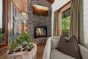 A cozy living room with a stone fireplace, a comfortable sofa, and a large window overlooking greenery. - Dogwood Dusk · New! Romantic Getaway – Hot Tub & Fireplace (Broken Bow)