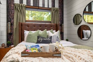 A neatly made bed with a 'stay awhile' pillow, a tray with a craft beer box, and a mug. - Dogwood Dusk · New! Romantic Getaway – Hot Tub & Fireplace (Broken Bow)