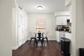 A compact kitchen with white cabinets, a dining table with chairs, and a window with blinds.