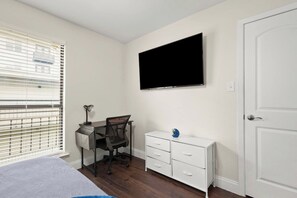 A bedroom with a bed, a desk with a chair, a TV mounted on the wall, and a white dresser.