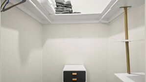 A corner closet with a white wall, a wooden floor, a small black and white storage unit, and a stack of folded towels on a shelf. - The Loop Loft | Stylish Stay Near Q2 Stadium (Austin)