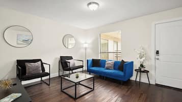 Uma sala de estar com um sofá azul, duas cadeiras pretas, uma mesa de centro e um espelho redondo na parede.