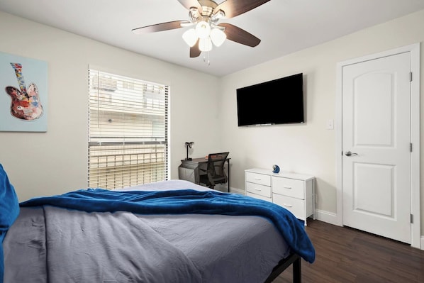 A bedroom with a bed, a desk, a chair, a ceiling fan, a TV, and a door.