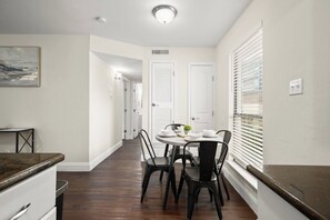 A narrow hallway leading to a dining area with a round table and chairs, white doors, and a window with blinds.