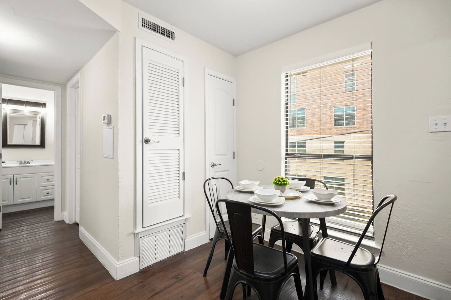 A room with a dining table set for four, a window with blinds, and a view of a brick building.