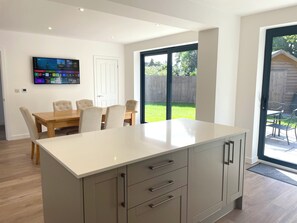 A modern kitchen with a large island, wooden dining table, and chairs. There is a TV mounted on the wall and a sliding glass door leading to a backyard. - Brand New detached 3 bedroom 2 bathroom house on the edge of Epping Forest (Coopersale)