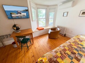 A room with a wooden floor, a bed with a quilt, a desk with a chair, a window seat, and a television displaying a boat on water.