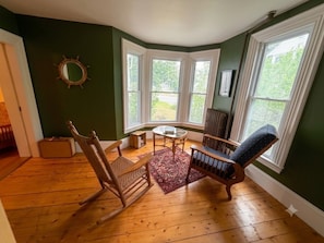 A room with a bay window, wooden flooring, a rocking chair, a blue upholstered chair, and a small table with a rug. - Towne Motel Suite #1 (Camden)