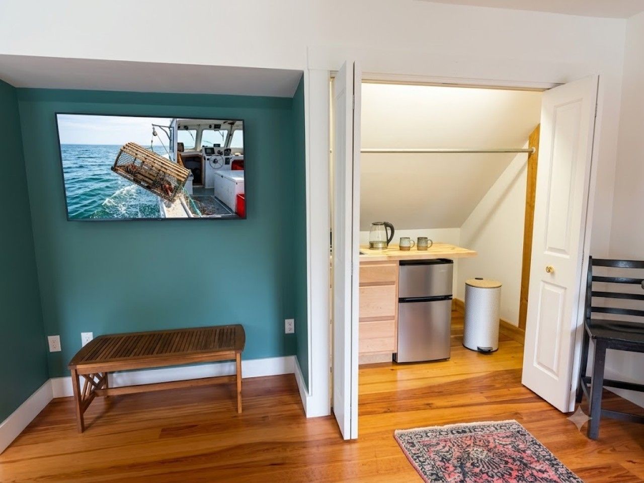 A room with a wooden bench, a kitchen area with a refrigerator, and a door leading to another room.
