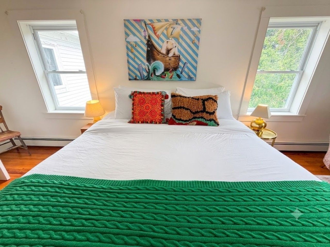 A bedroom with a large bed, two windows, a painting on the wall, and a green blanket.