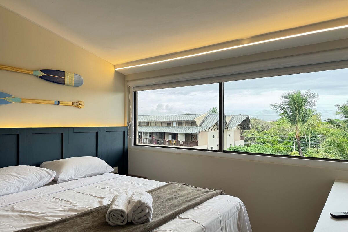 A hotel room with a bed, a window with a view of buildings and greenery, and a wall-mounted telephone.