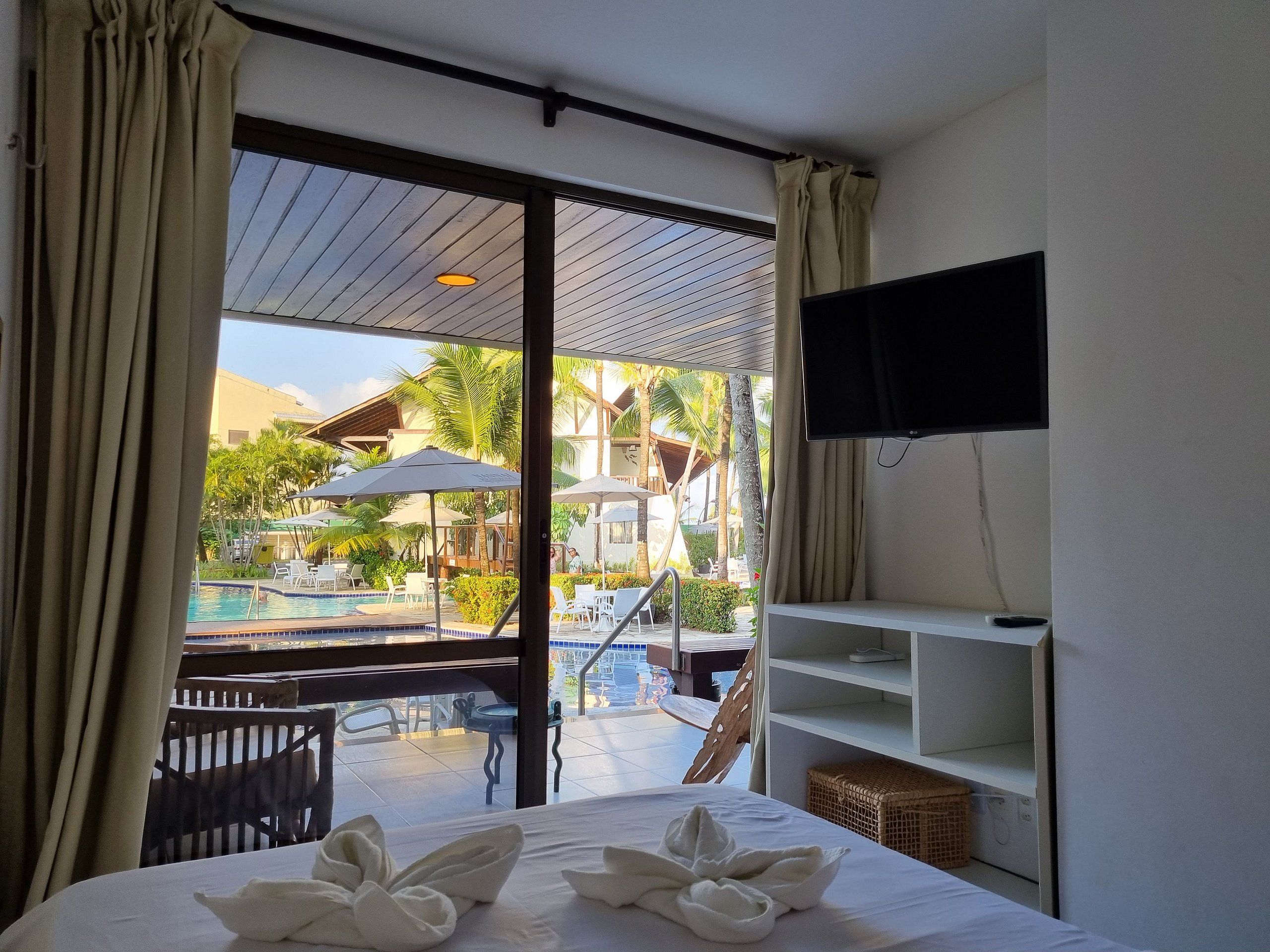 A hotel room with a bed, a TV mounted on the wall, and a view of a pool and outdoor seating area.