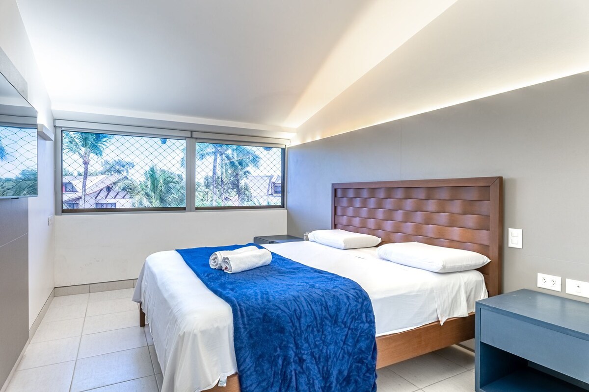 A modern hotel room with a large bed, a nightstand, and a window with a view of palm trees.