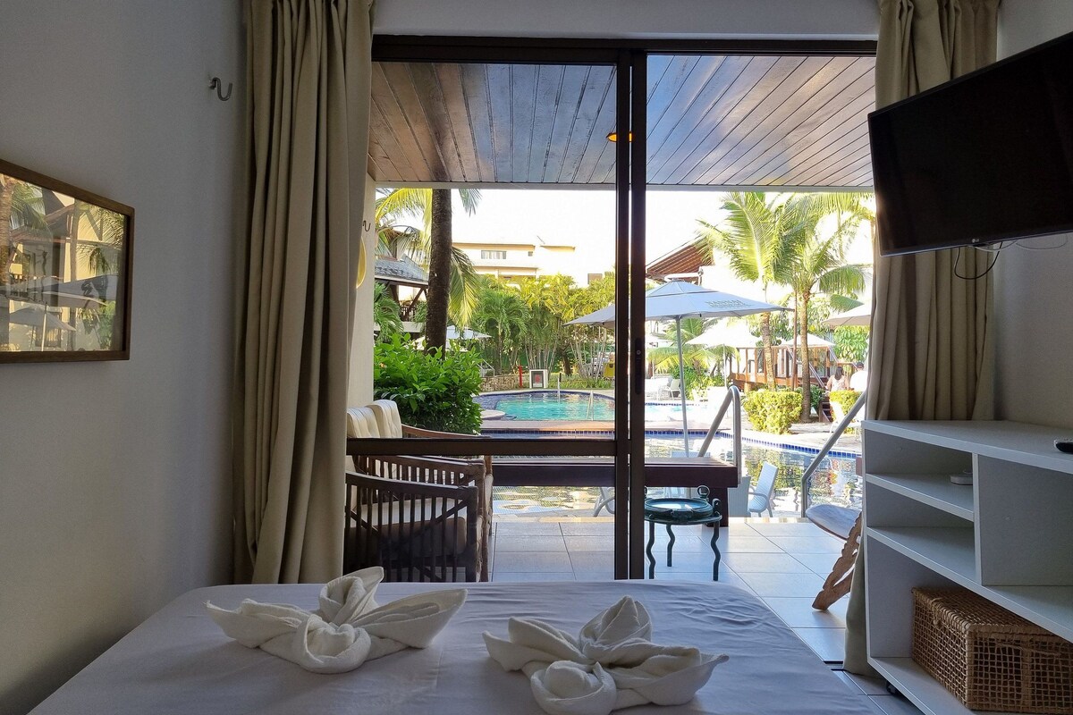 A hotel room with a bed, a television, a window with curtains, a view of a pool and outdoor seating, and a framed picture on the wall.