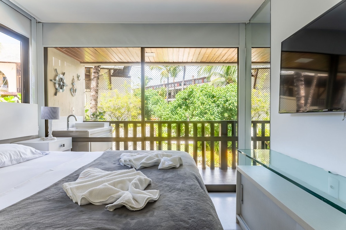 A modern bedroom with a large bed, a bedside table, a lamp, and a view of a balcony with a wooden railing.