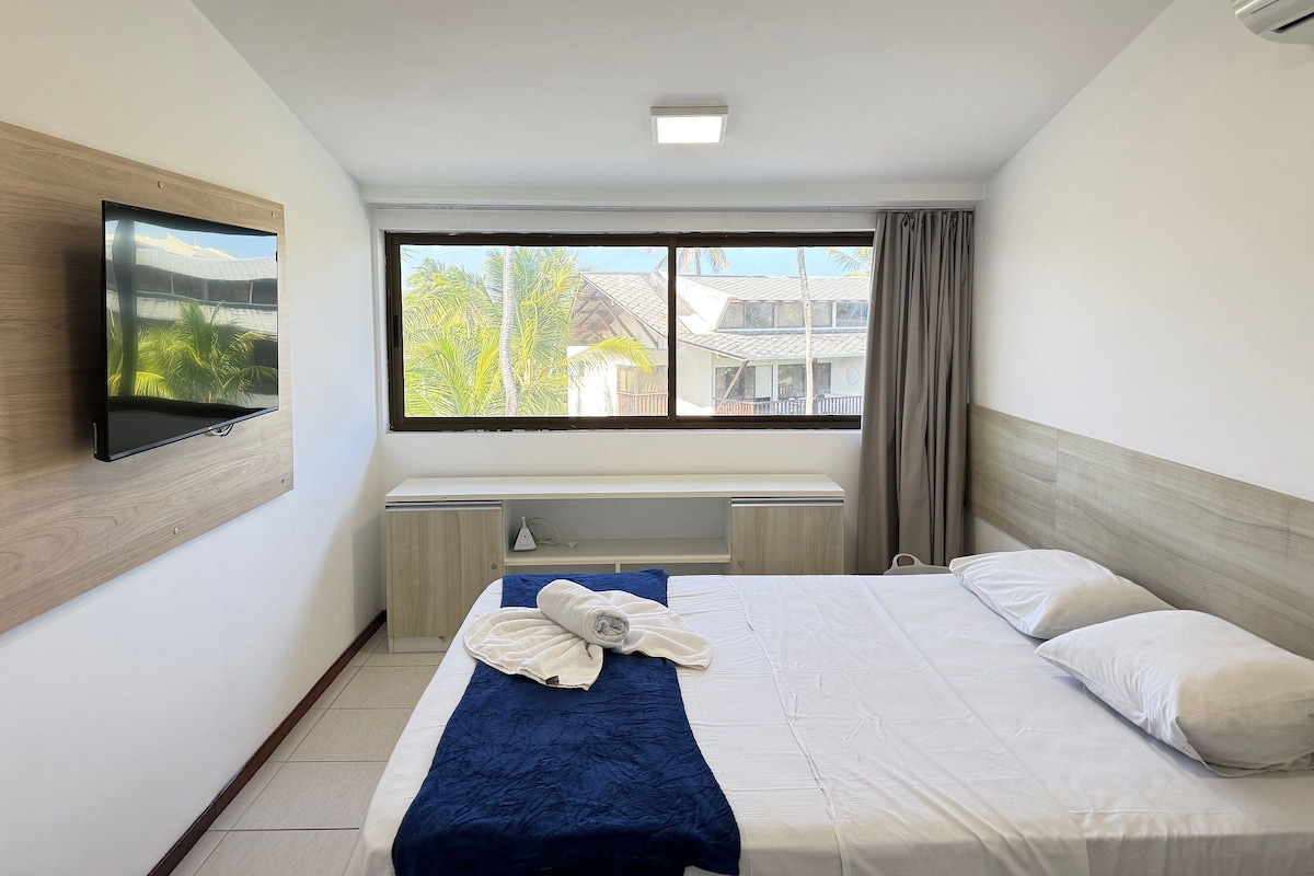A hotel room with a bed, a desk, a flat-screen TV, and a window with a view of palm trees and a building.