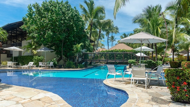 A swimming pool with lounge chairs and umbrellas, surrounded by palm trees and a stone deck. - Nannai Residence Muro Alto Beira Mar (N. Sra. do Ó)