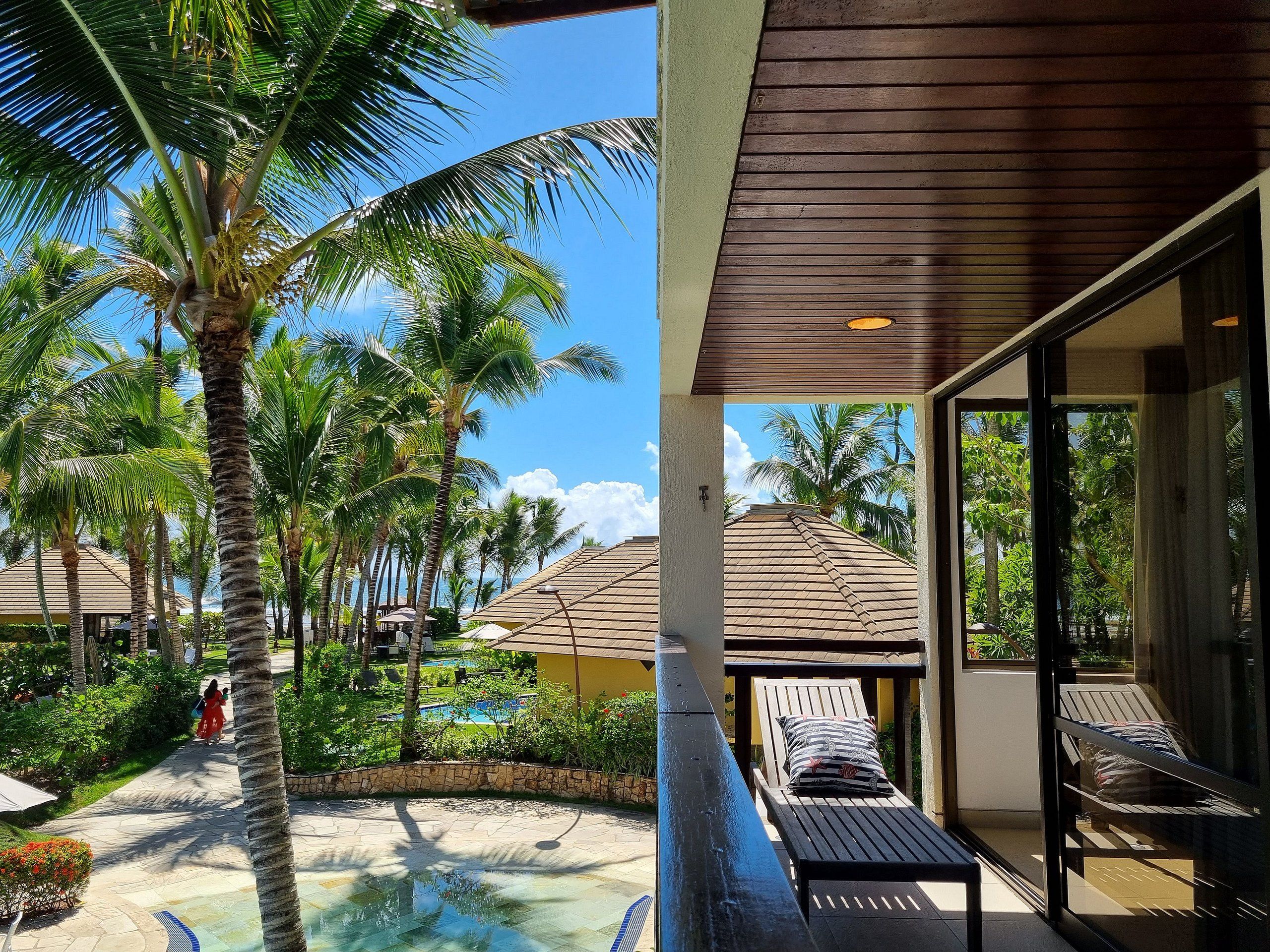 Un espace aménagé au bord de la piscine, avec des chaises longues, un palmier et une vue dégagée sur la piscine et les autres bâtiments.