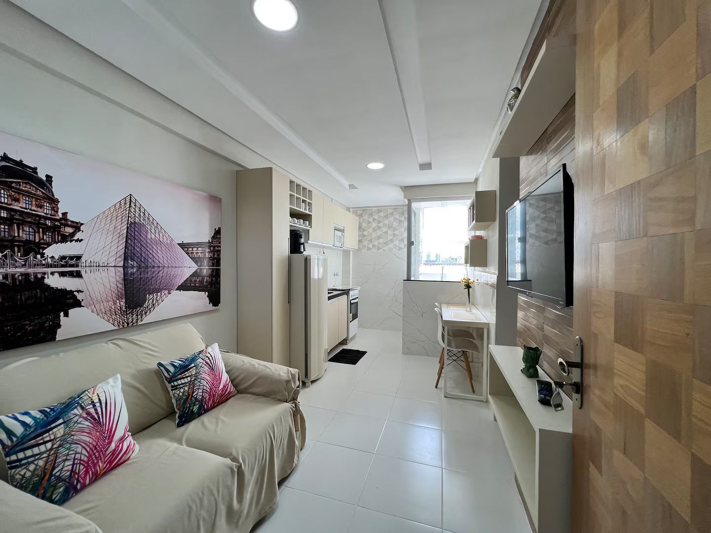 Una sala de estar moderna con un sofá, un televisor montado en la pared y una zona de cocina con refrigerador y microondas.