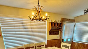 Uma sala de jantar com mesa, cadeiras, um candelabro e uma área de cozinha com geladeira e armários.