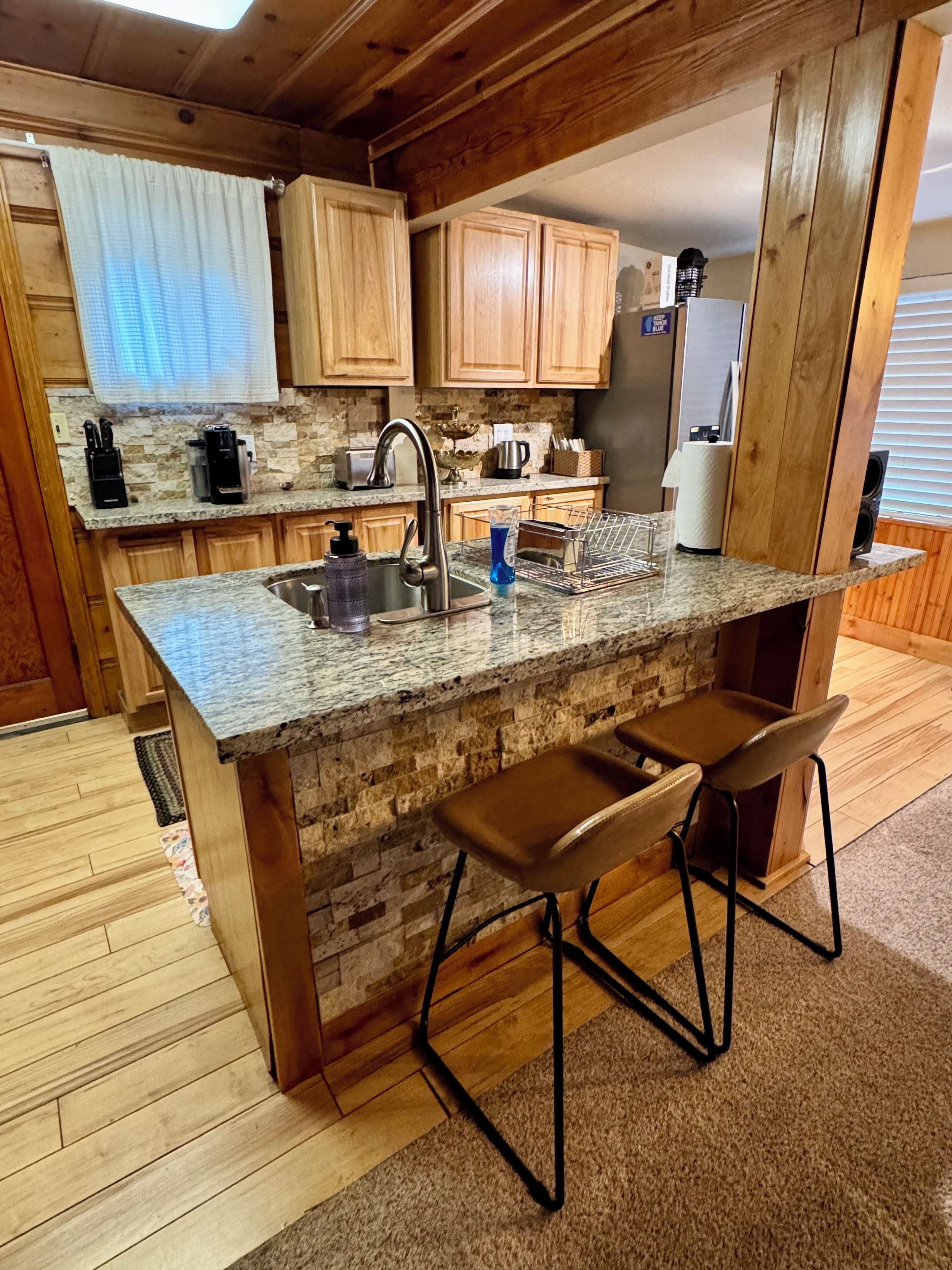 A kitchen with a granite countertop, wooden cabinets, a stainless steel sink, and a bar stool.