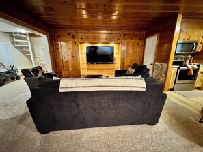 A cozy living room with a dark-colored sofa, a flat-screen TV, wooden paneling, a staircase, and a kitchen area with modern appliances. - Entire cabin in South Lake Tahoe, California (South Lake Tahoe)