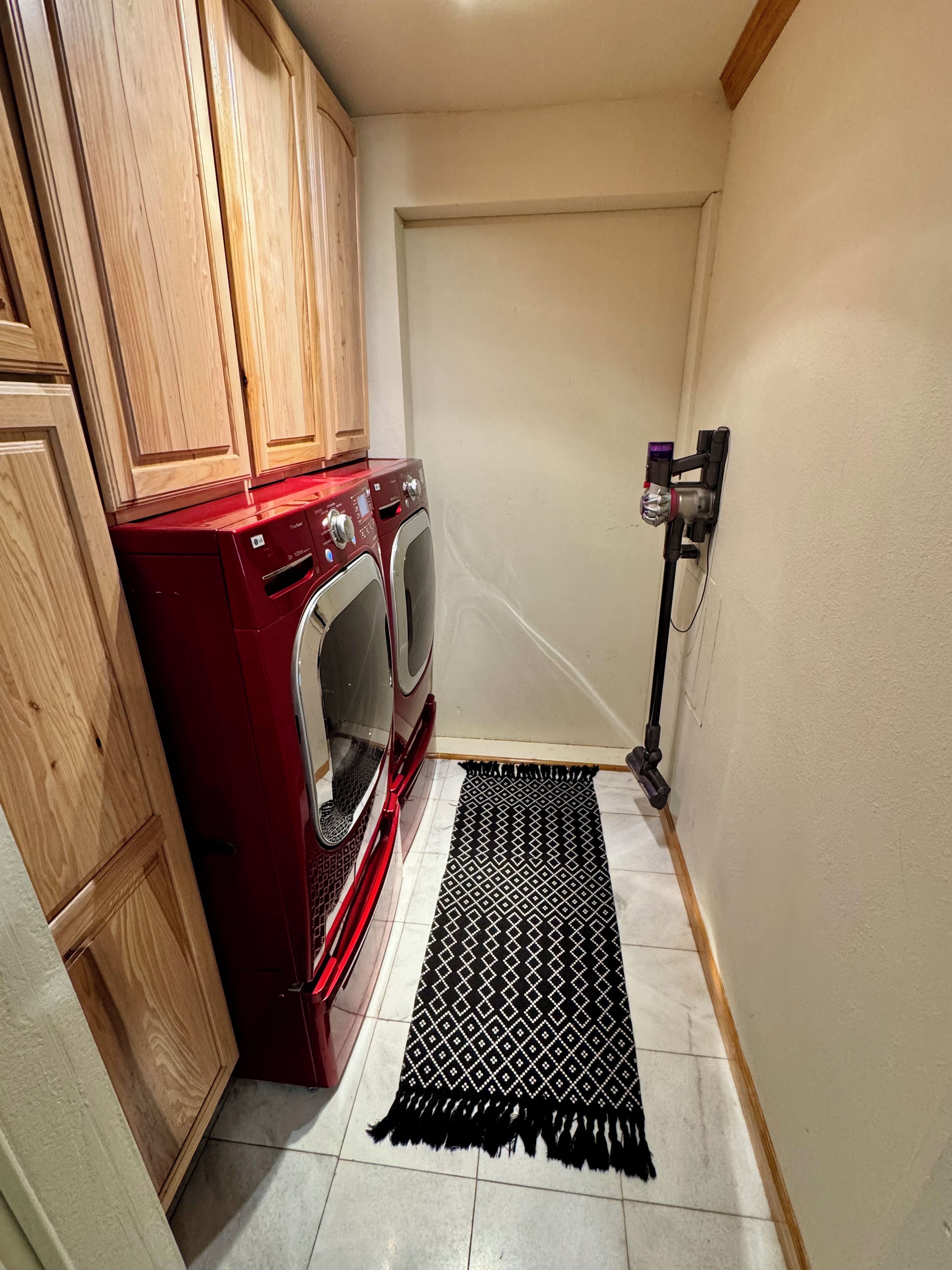 Een wasruimte met een rode wasmachine, houten kastjes, een zwart tapijt met patroon en een stofzuiger aan de muur.