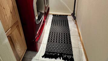 Een wasruimte met een rode wasmachine, houten kastjes, een zwart tapijt met patroon en een stofzuiger aan de muur.