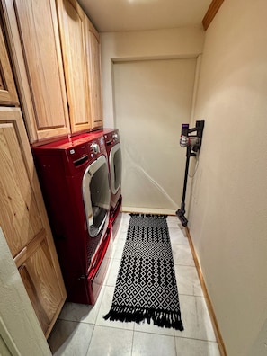 A laundry area with a red washing machine, wooden cabinets, a black patterned rug, and a wall-mounted vacuum cleaner.