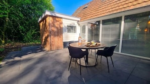 Un patio exterior con una mesa redonda y sillas, una estructura de madera y una casa con techo de tejas y ventanas grandes.