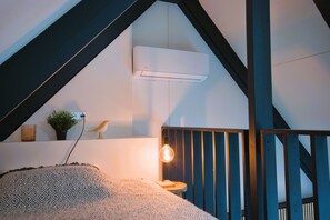 Una habitación acogedora con techo inclinado, aire acondicionado instalado, una mesita con lámpara y una escultura decorativa de un pájaro.