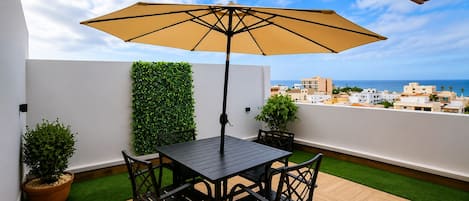 A terrace with a table and chairs, a large umbrella, and a view of the city and sea.