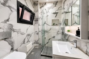 Un baño moderno con cabina de ducha de vidrio, un lavabo blanco y una zona de ducha con azulejos de mármol.