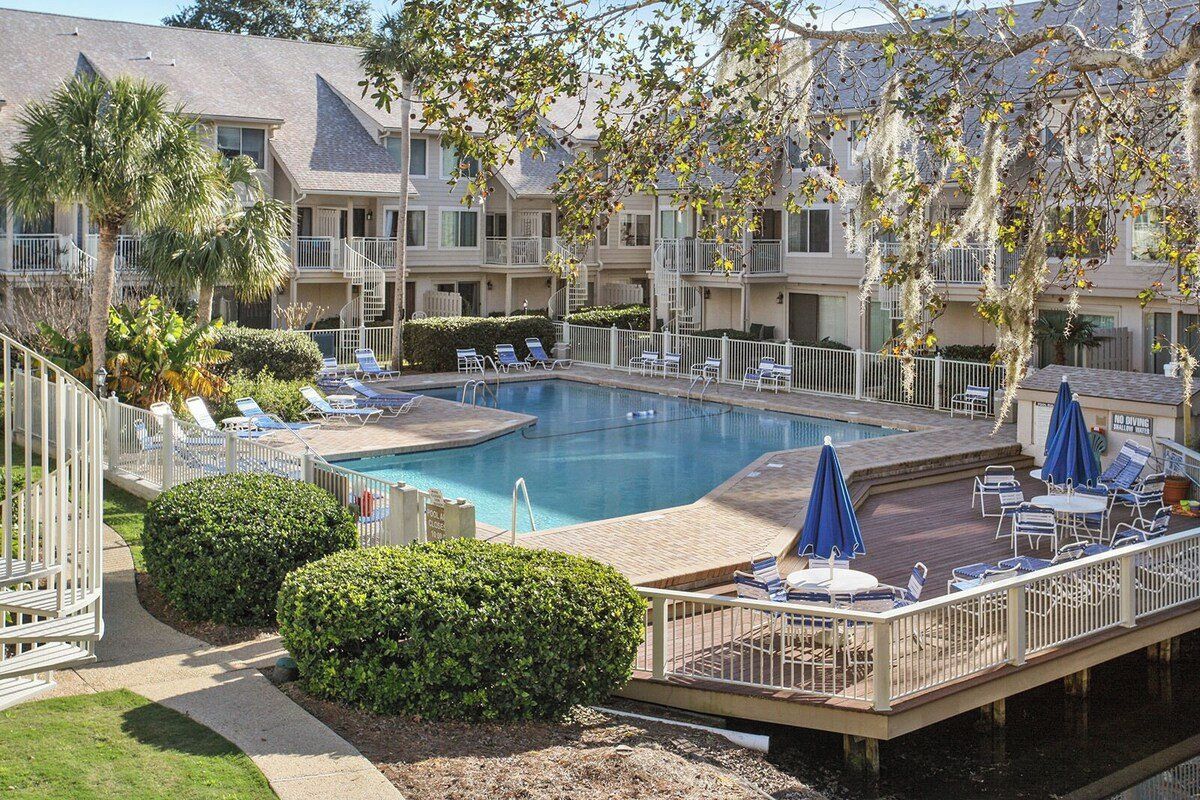 A hotel pool area with lounge chairs and umbrellas, surrounded by a deck and a white fence.