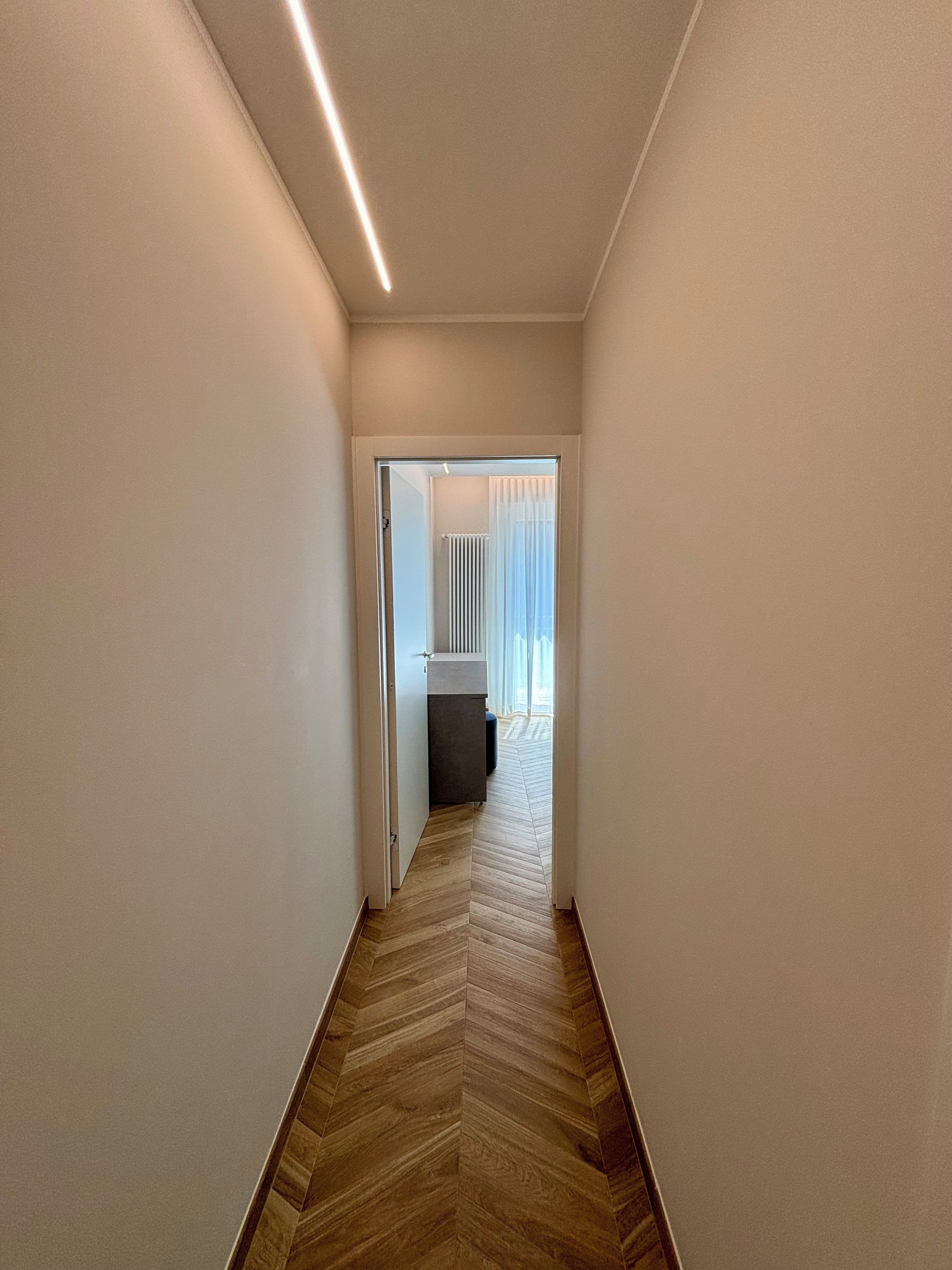 A narrow hallway with a herringbone floor pattern, leading to a room with a radiator and a window with sheer curtains.
