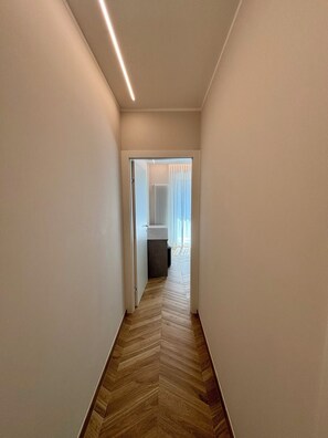 A narrow hallway with a herringbone floor pattern, leading to a room with a radiator and a window with sheer curtains. - Luxurious new apartment with a terrace overlooking the lake on the Sarnico lakeshore. (Sarnico)