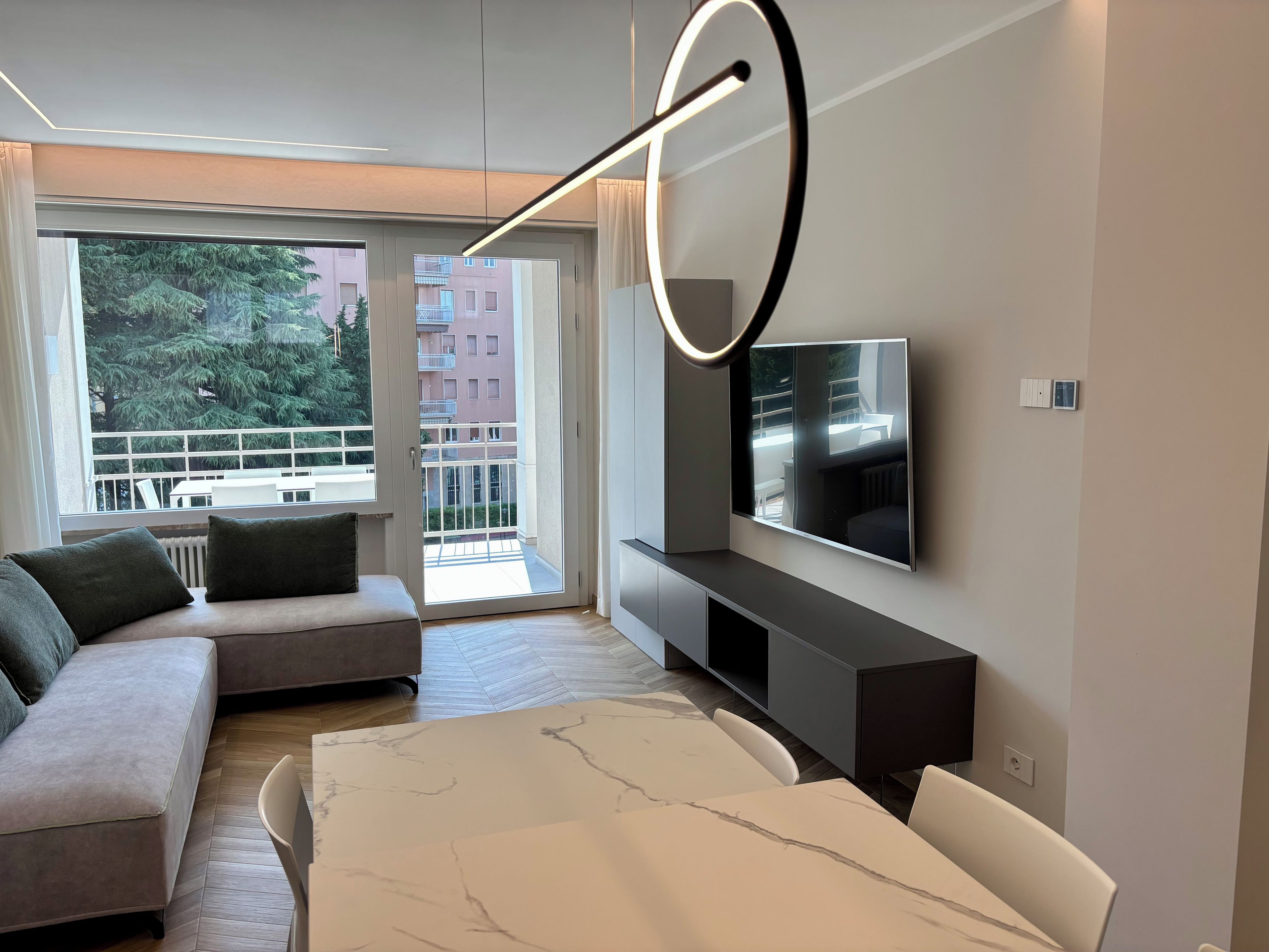 A modern living room with a sofa, a large flat-screen TV, and a balcony with a view of buildings and trees.