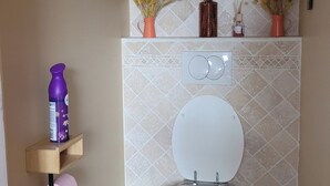 A bathroom with a toilet, a hand dryer, a trash bin, a toilet brush, and a shelf with a bottle and a roll of toilet paper. - LE CLOS DES VOYAGEURS Bed and Breakfast ALL THE COMFORTS (Saint-Christophe-de-Double)